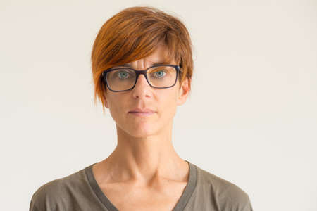 Waist up portrait of mature woman with red hairs, green eyes, eye glasses and serious facial expression, standing against the wall. Natural soft daylight, natural skin, neutral background.の写真素材