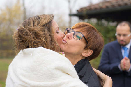 Best friend congratulating, hugging and kissing the bride during the wedding ceremony. Groom out of focus in the background. Shot outdoors, real people.の写真素材