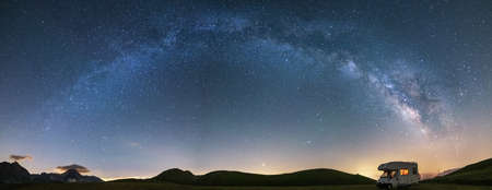 Panoramic night sky over Campo Imperatore highlands, Abruzzo, Italy. The Milky Way galaxy arc and stars over illuminated camper van. Camping freedom in unique hills landscape.の写真素材