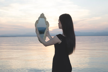 Woman in black dress with mirror in front of the sea: reflections between reality and introspectionの写真素材