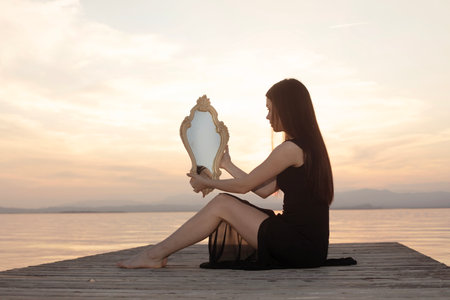 elegant woman with mirror mirrors her soul in front of a sunset landscape on the sea, concept of identityの写真素材