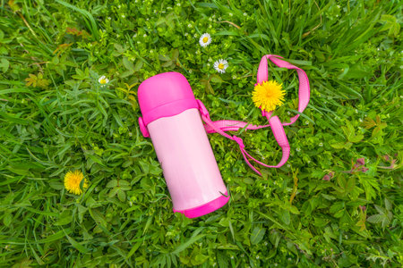 Children's pink flask on green grass. Reusable thermo bottle for drinks, water, tea. Flask for resting and relax outdoorsの写真素材