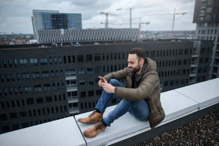 Smiling and bearded hipster guy looks at the social network in the smartphone, on the roof of a tall building.の写真素材
