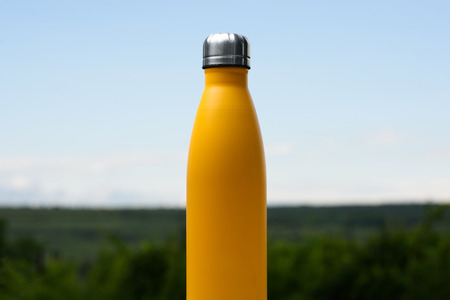Stainless bottle for water, tea or coffee. Sky and forest on background. On the glass desk.の写真素材