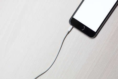 Smartphone with mockup screen and cable from charging on white wooden table background