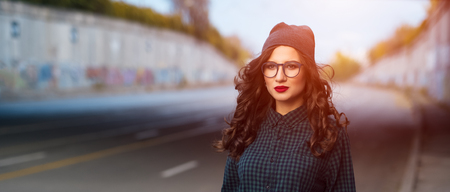 Urban young curly girl, standing in street at sunset, wearing glasses and dressed with shirt and hat.の写真素材
