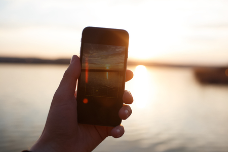 Male hand holding smartphone and taking the photo of sunset.の写真素材