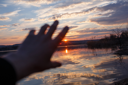 Male hand is touching the sunset over lake.の写真素材
