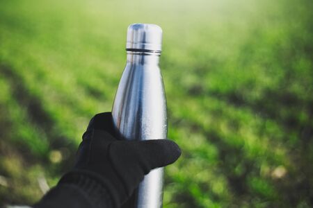 Close-up of male hand dressed in glove holding reusable steel thermo water bottle on background of blurred green grass.	の写真素材