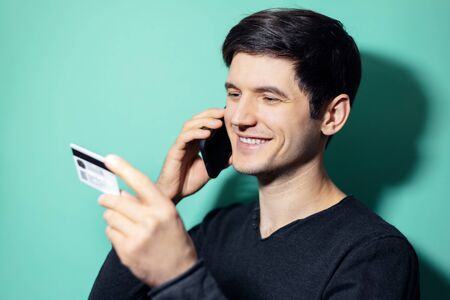 Studio portrait of young smiling man talking on smartphone and looking at his credit card in hand on background of aqua menthe color.の写真素材