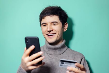 Studio portrait of young happy man with smartphone and credit card in hand on background of aqua...