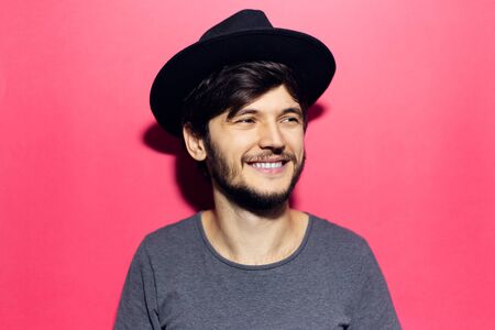 Portrait of young smiling guy, wearing black hat and grey shirt, isolated on pink background.