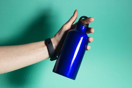 Close-up of female hand holding a reusable, aluminium thermal water bottle of phantom blue color,...