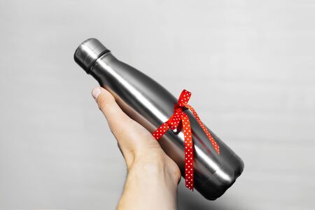 Close-up of male hand holding a eco water thermo bottle of steel with red ribbon, ecologic gift for any events. Isolated on white background. Say no to plastic when you have another choice.の写真素材
