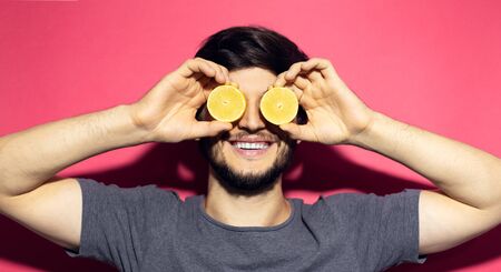 Close-up portrait of young happy man holding lemons on eyes, isolated on pink coral background.