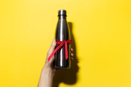 Close-up of male hand holding a eco water thermo bottle of steel with red ribbon, ecologic gift for any events. Isolated on yellow background with shadows.の写真素材