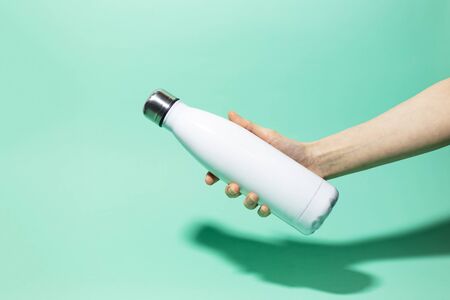 Close-up of female hand holding white reusable steel stainless thermo water bottle isolated on background of aqua menthe color. Plastic free.の写真素材