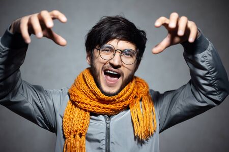 Portrait of young angry man making cat claws gesture, wearing glasses and orange scarf, background...