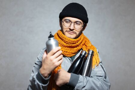 Portrait of young man, holding steel and aluminium thermo eco water bottles, wearing glasses, orange...