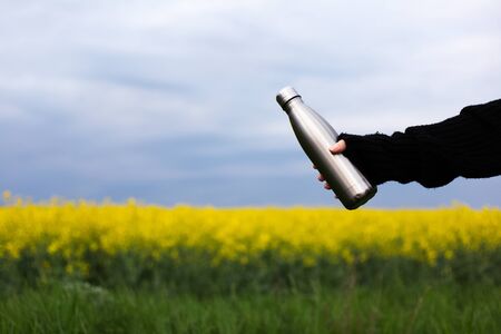 Stainless vacuum flask bottle in hand, on background of rapeseed fieldの写真素材