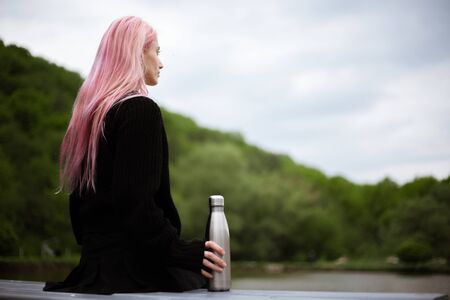 Portrait of young girl with pink hair sitting in park with thermo bottle in handの写真素材