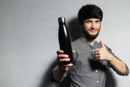 Portrait of self-confident bearded guy, showing thumbs up for steel ecologic black bottle for water. Say no to plastic. Textured background of grey.の写真素材