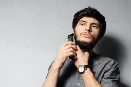 Portrait of young bearded guy shaving beard with trimmer. On textured background of grey.