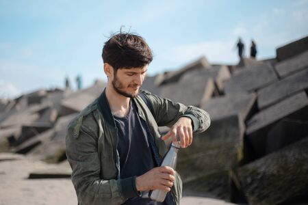 Portrait of young man with backpack, using reusable steel thermo water bottle, outdoor on background of square stones.の写真素材