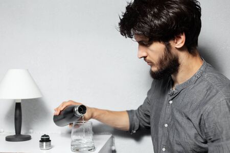 Portrait of young bearded guy with disheveled hair, pours water in glass from steel thermo bottle, on background of grey.の写真素材