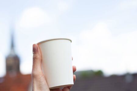 Close-up of female hand hold white paper glass with mockup on background of sky.