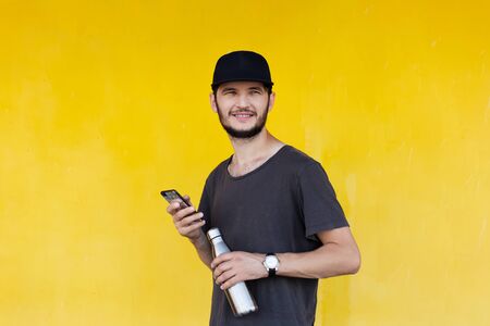 Portrait of young smiling guy with thermo bottle for water and smartphone in hands. On background of...