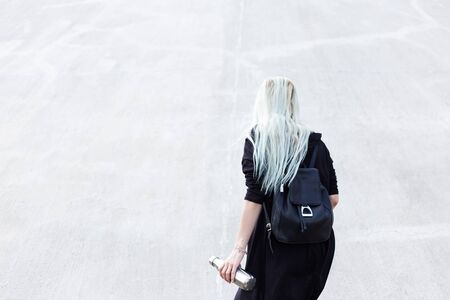 Back view of young blonde girl, dressed in black with backpack, holding steel thermo bottle in hand. On background of textured light grey wall.の写真素材