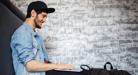 Portrait of young man in denim jacket and black cap sitting on the floor, working on laptop, using smartphone, black cup of coffee.の写真素材