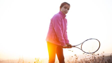 Portrait of teenage girl is training tennis skill outside court, in field at sunset. Wearing pink sweater and holding tennis racket.の写真素材