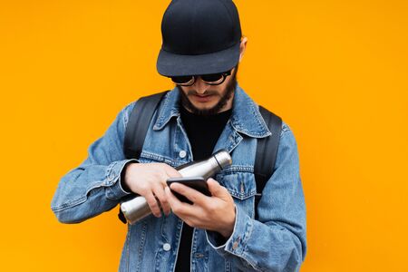 Portrait of young self-confident hipster man on orange background. Using smartphone with steel thermo eco bottle for water in another hand.の写真素材