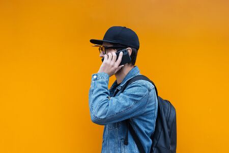 Portrait of young hipster talking on smartphone on orange background. Wearing jeans jacket, cap,...