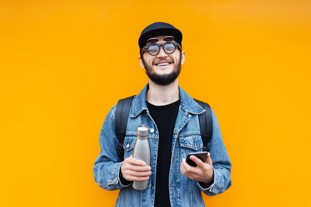 Portrait of young cheerful hipster man on orange background. Holding steel thermo eco bottle for...