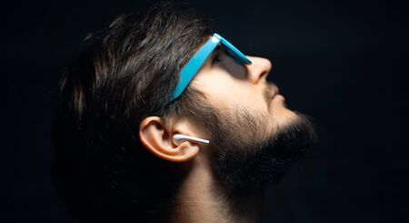 Close-up of profile portrait of young guy with earbuds, looking up, wearing cyan sunglasses,...