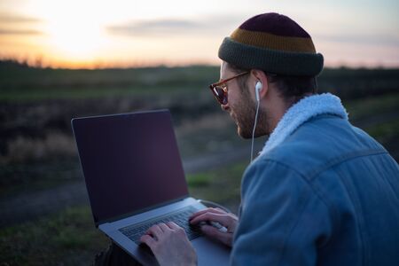 Millennial hipster man working on laptop with earphones in field at sunset.