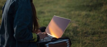 Portrait of young hipster girl in denim jacket, using laptop with earphones in backpack on background of sunset in field.の写真素材