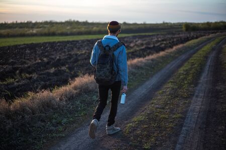 Back view of young guy with backpack walking on village road with aluminum reusable water bottle in...