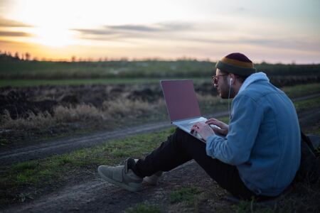 Millennial hipster man working on laptop with earphones in field at sunset.の写真素材