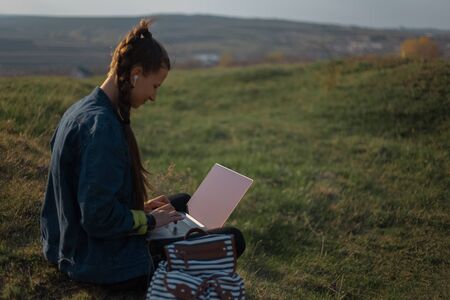 Portrait of young hipster girl in denim jacket, using laptop with earphones in backpack on...
