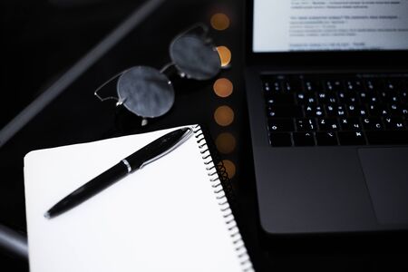 Top view of modern, black work table with laptop, notebook, pen and mirror sunglasses.
