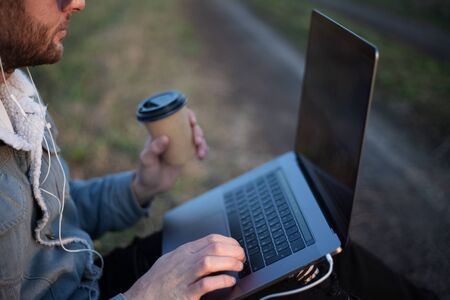 Close-up of young man sitting with laptop on legs and cup of coffee in hand.の写真素材