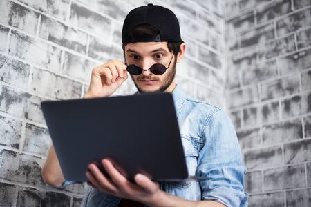Portrait of young man looking in laptop with sunglasses and black cap on the background of grey...