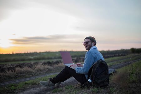 Millennial hipster man working on laptop with earphones in field at sunset.の写真素材