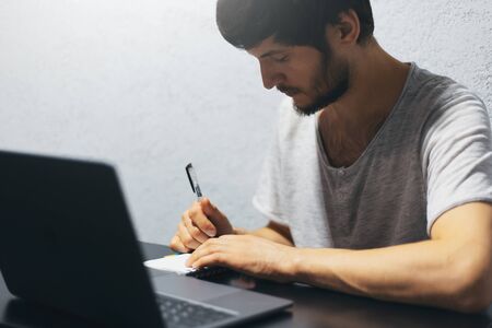 Portrait of young man writing in notebook, working with laptop.の写真素材