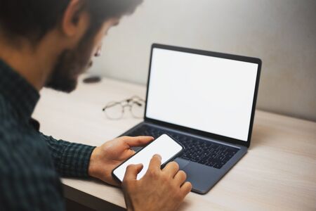 Close-up of male hands, touching screen of smartphone on background of laptop on desk, with mockup.