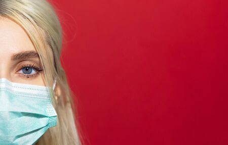 Close-up portrait of young blonde girl wearing medical flu mask on background of red color with copy...